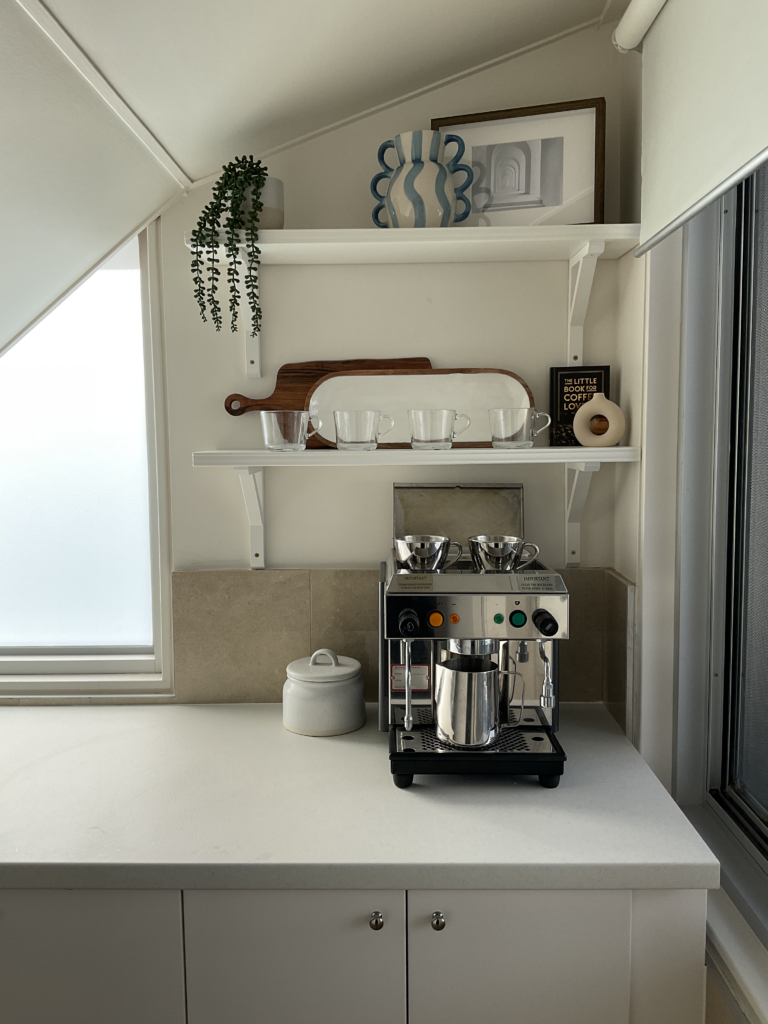 Coffee bar area of the pantry. Includes lower cabinets with a benchtop for a coffee machine and then two open shelves which are styled with coffee mugs.