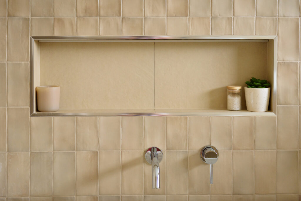 Closeup image of the bathtub niche showing feature wall tile with the large format tile inside the niche. Styling includes a candle, bath salts and a small plant. Underneath the niche is chrome tapware from ABI Interiors