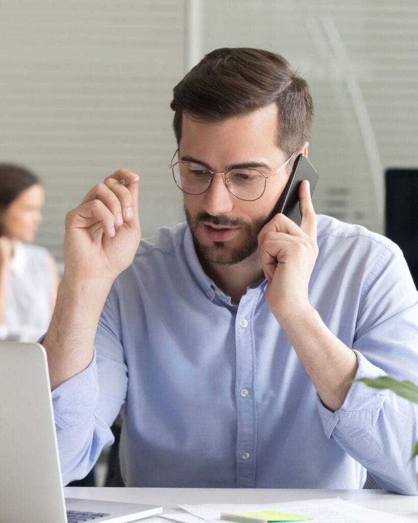 Stock image of a man in glasses talking in a phone, in front of a laptop, to illustrate the recommendation to have one main contact