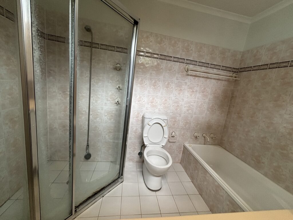 Second before photo of the bathroom in this semi renovation with a shower, toilet and bath with outdated pink tiles